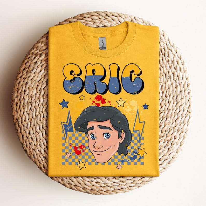 May include: Mustard yellow t-shirt featuring the name "ERIC" in large, retro-style blue and yellow lettering. The shirt also displays a cartoon illustration of a character with black hair and blue eyes, along with stars and lightning bolts.