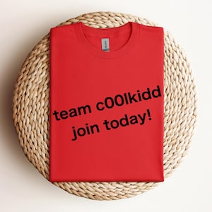 May include: A folded red t-shirt with the text "team c00lkidd join today!" in black. The shirt is on a round, woven, light brown surface.
