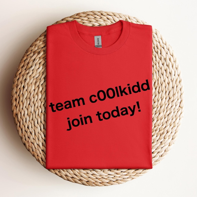 May include: A folded red t-shirt with the text "team c00lkidd join today!" in black. The shirt is on a round, woven, light brown surface.