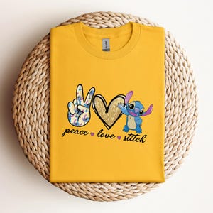 以下が含まれることがあります： リロ＆スティッチのスティッチが描かれた黄色のTシャツ。平和のサイン、ハート、そして「peace love stitch」の文字が書かれています。