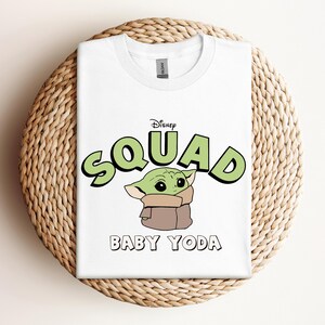May include: White t-shirt featuring the text "Disney Squad" in green with a black outline, and a cartoon image of Baby Yoda. The shirt is folded on a woven, circular surface.