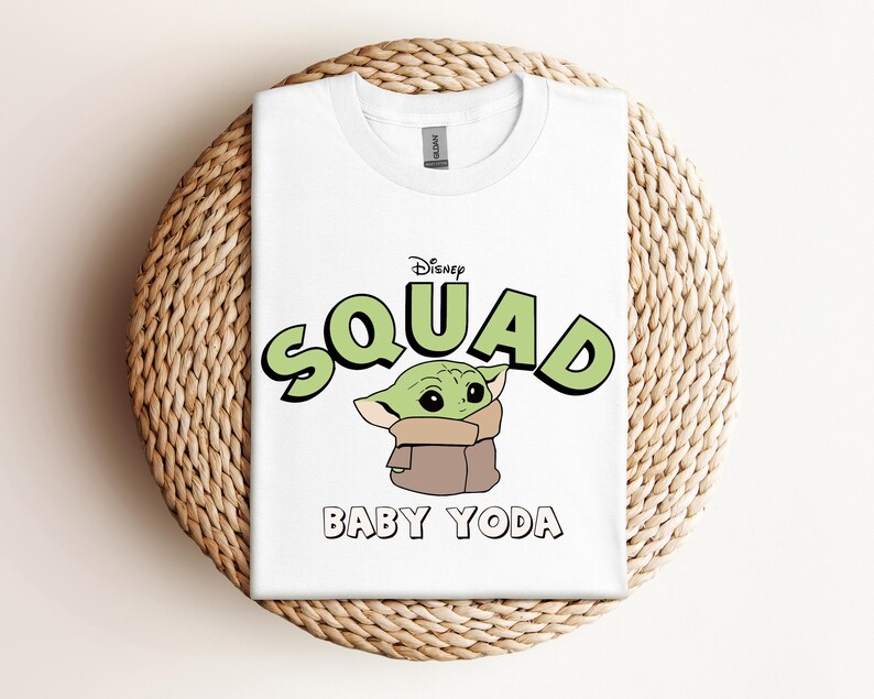 May include: White t-shirt featuring the text "Disney Squad" in green with a black outline, and a cartoon image of Baby Yoda. The shirt is folded on a woven, circular surface.
