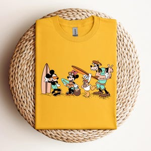 May include: Yellow t-shirt with a graphic of Mickey Mouse, Minnie Mouse, Donald Duck, and Goofy on a beach with a surfboard. The characters are all wearing summer clothes and are smiling.