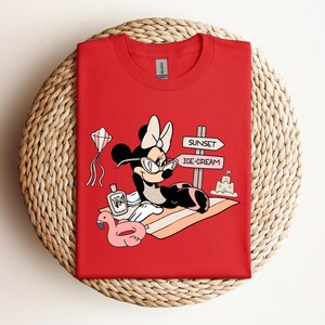 K&ouml;nnte beinhalten: Rotes T-Shirt mit einem Cartoon-Bild von Minnie Mouse, die eine Sonnenbrille und eine Schleife tr&auml;gt und sich auf einem Strandtuch mit einem rosafarbenen Flamingo-Schwimmtier, einer Flasche Sonnencreme und einem Schild mit der Aufschrift "Sunset" und "Ice-Cream" entspannt.