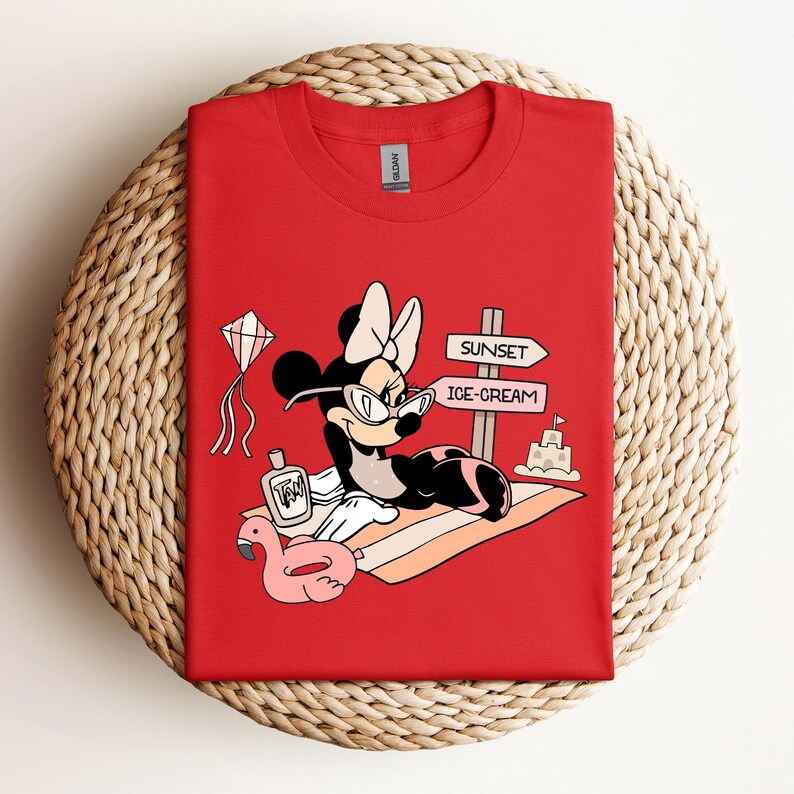 K&ouml;nnte beinhalten: Rotes T-Shirt mit einem Cartoon-Bild von Minnie Mouse, die eine Sonnenbrille und eine Schleife tr&auml;gt und sich auf einem Strandtuch mit einem rosafarbenen Flamingo-Schwimmtier, einer Flasche Sonnencreme und einem Schild mit der Aufschrift "Sunset" und "Ice-Cream" entspannt.