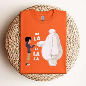 K&ouml;nnte beinhalten: Orangefarbenes T-Shirt mit einem wei&szlig;en Cartoon-Roboter und einem Jungencharakter. Der Text "BA LA LA LA LA" ist auf dem T-Shirt aufgedruckt.