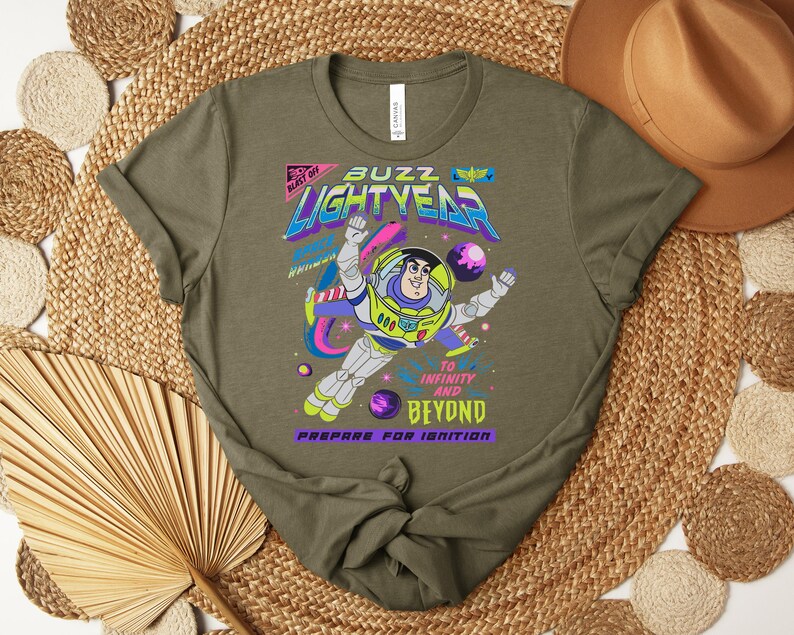 Puede incluir: Camiseta verde oliva con un gr&aacute;fico de Buzz Lightyear en un traje espacial, con las palabras "Buzz Lightyear" y "To Infinity and Beyond". El dise&ntilde;o incluye planetas y un tema espacial colorido.