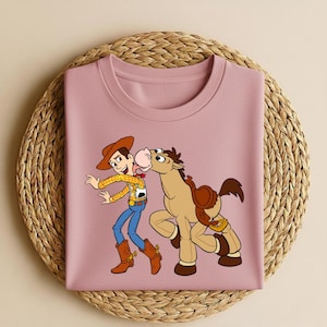 Peut inclure: Un t-shirt rose clair avec une illustration de dessin anim&eacute; de Woody et Bullseye de Toy Story. Woody porte un chapeau de cowboy, une chemise jaune, un jean bleu et des bottes marron. Bullseye est un cheval marron avec une selle.