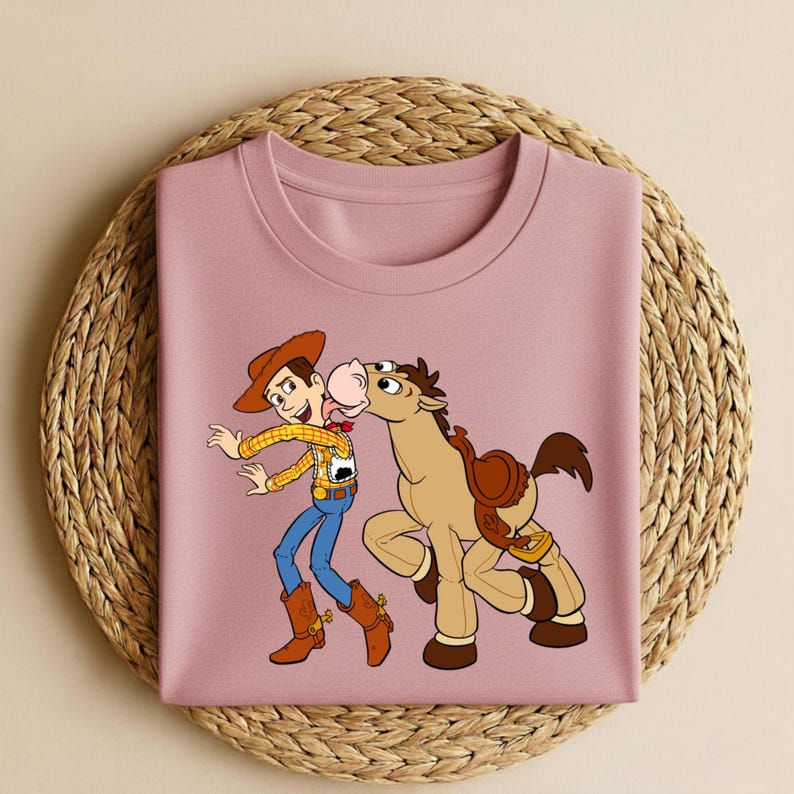 Peut inclure: Un t-shirt rose clair avec une illustration de dessin anim&eacute; de Woody et Bullseye de Toy Story. Woody porte un chapeau de cowboy, une chemise jaune, un jean bleu et des bottes marron. Bullseye est un cheval marron avec une selle.