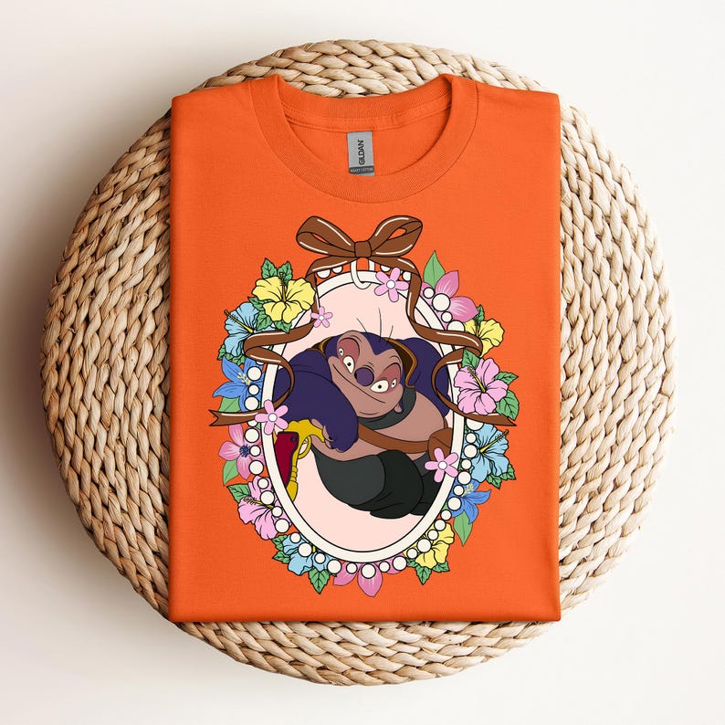 Puede incluir: Camiseta naranja con un personaje de dibujos animados dentro de un marco ovalado. El marco est&aacute; decorado con flores y una cinta marr&oacute;n. El personaje es una criatura azul y morada con una expresi&oacute;n traviesa.