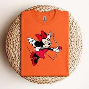 Puede incluir: Camiseta naranja con una imagen de dibujos animados de Minnie Mouse vestida de Cupido, sosteniendo un arco y una flecha con una punta de flecha en forma de corazón.