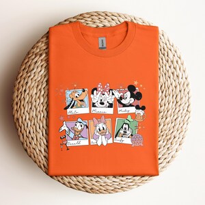May include: An orange crew neck t-shirt featuring a graphic print of six individual polaroid-style frames. Each frame contains a different Disney character illustration with their name written below, including Pluto, Minnie, Mickey, Donald, Daisy, and Goofy.