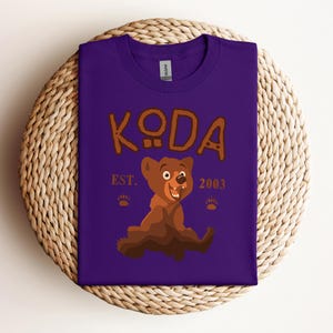 May include: Purple t-shirt with a brown cartoon bear and the text "KODA EST. 2003".