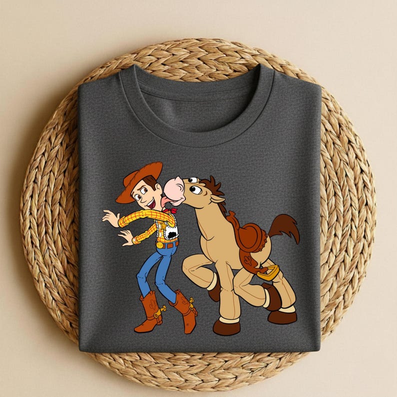 Peut inclure: T-shirt gris fonc&eacute; avec une illustration de Woody et Bullseye de Toy Story. Woody porte un chapeau de cowboy, une chemise &agrave; carreaux et un jean bleu. Bullseye est un cheval marron avec une selle. Le t-shirt est sur un tapis tiss&eacute;.