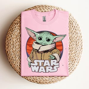 Op de afbeelding: Lichtroze T-shirt met een afbeelding van een groen buitenaards personage uit Star Wars, met grote oren en een bruine jas. Het personage staat tegen een rode en oranje cirkelvormige achtergrond, met de woorden "STAR WARS" in het wit eronder.