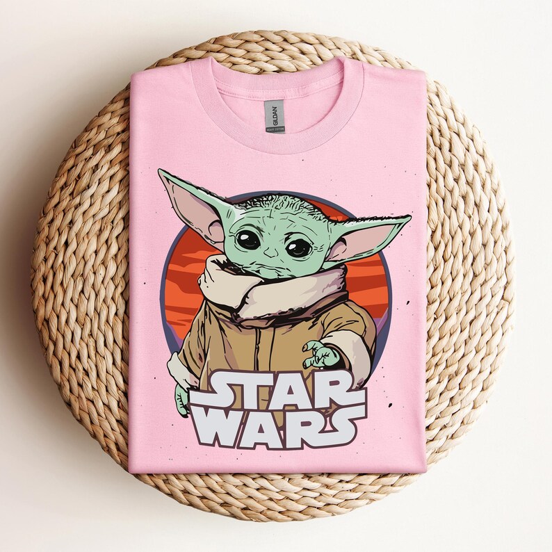 Op de afbeelding: Lichtroze T-shirt met een afbeelding van een groen buitenaards personage uit Star Wars, met grote oren en een bruine jas. Het personage staat tegen een rode en oranje cirkelvormige achtergrond, met de woorden "STAR WARS" in het wit eronder.