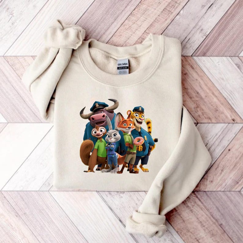 Puede incluir: Sudadera de color crema con un gr&aacute;fico colorido de personajes de la pel&iacute;cula animada "Zootr&oacute;polis". El dise&ntilde;o incluye varios personajes de animales con uniformes de polic&iacute;a y ropa informal. La sudadera est&aacute; doblada sobre una superficie de madera.