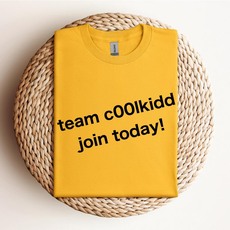 May include: Mustard yellow t-shirt with the text "team c00lkidd join today!" in black. The shirt is folded and placed on a woven, circular surface. The Gildan brand label is visible on the collar.