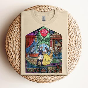 May include: Beige t-shirt featuring a colorful stained-glass design. The design depicts a couple dancing, a red rose, and various characters. The shirt is folded on a woven, circular surface.