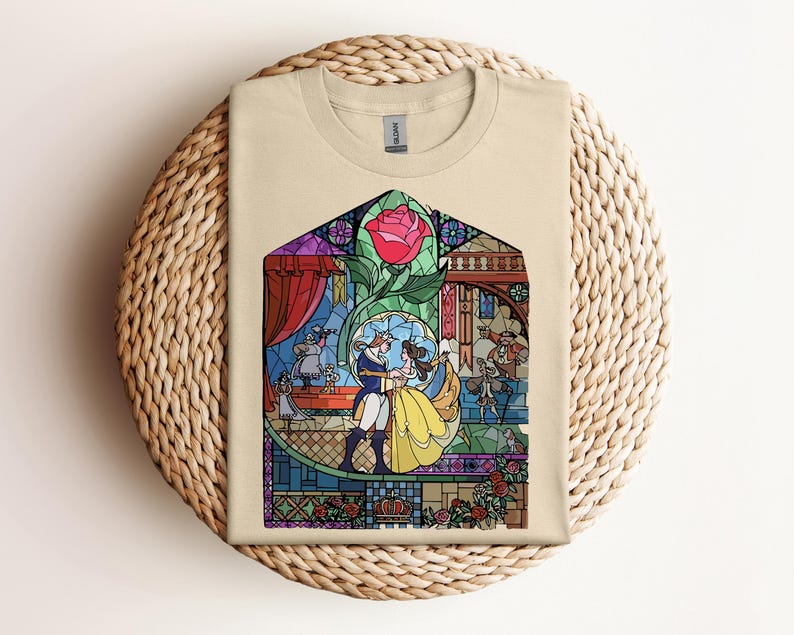 May include: Beige t-shirt featuring a colorful stained-glass design. The design depicts a couple dancing, a red rose, and various characters. The shirt is folded on a woven, circular surface.