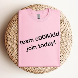 May include: A light pink t-shirt with the text "team c00lkidd join today!" in black. The shirt is folded and placed on a round, woven, beige placemat. The shirt has a crew neck and short sleeves.