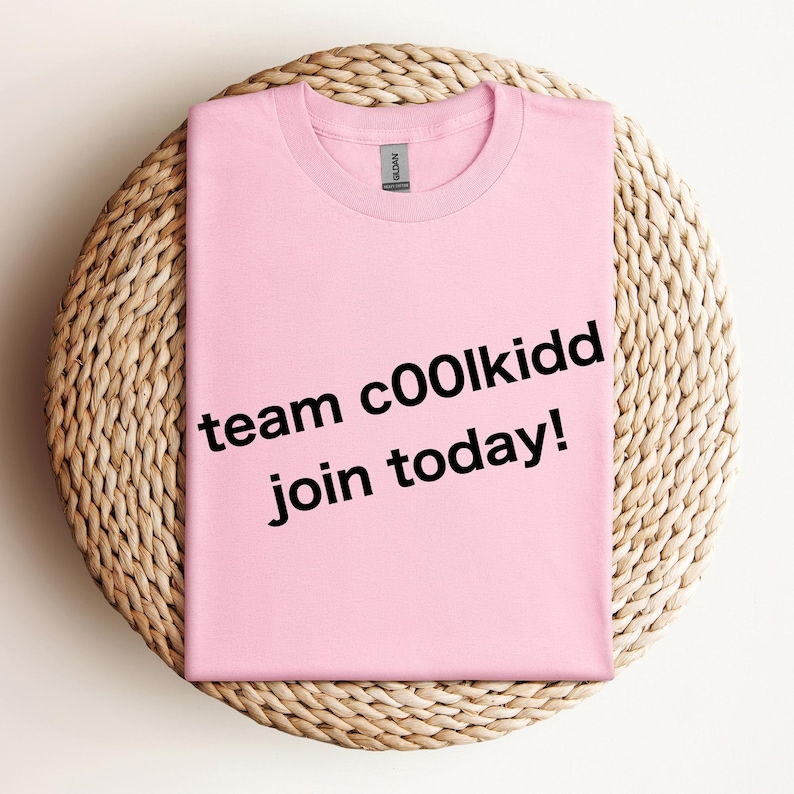 May include: A light pink t-shirt with the text "team c00lkidd join today!" in black. The shirt is folded and placed on a round, woven, beige placemat. The shirt has a crew neck and short sleeves.