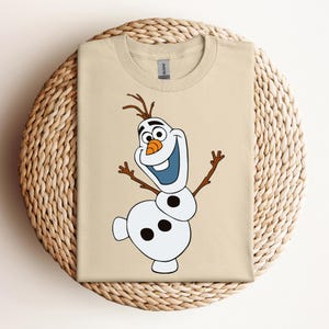 Pode incluir: Uma camiseta bege com uma imagem de desenho animado de Olaf, o boneco de neve do filme Frozen. Olaf est&aacute; sorrindo com os bra&ccedil;os estendidos e tem um nariz de cenoura.