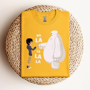 K&ouml;nnte beinhalten: Ein gelbes T-Shirt mit einem wei&szlig;en Roboter und einer Comicfigur. Der Text "BA LA LA LA" ist auf dem T-Shirt aufgedruckt.