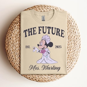Puede incluir: Camiseta beige con Minnie Mouse de dibujos animados con un vestido de novia, sosteniendo flores y un anillo. El texto "THE FUTURE" está encima de la imagen, con "EST. 2025" y "Mrs. Warling" debajo. La camiseta se muestra en una estera tejida.