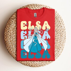 Op de afbeelding: Rood T-shirt met het woord "ELSA" in een retro lettertype, en een afbeelding van Elsa uit Frozen. Het ontwerp bevat sneeuwvlokken en herfstbladeren. Het T-shirt wordt getoond op een geweven, cirkelvormig oppervlak.