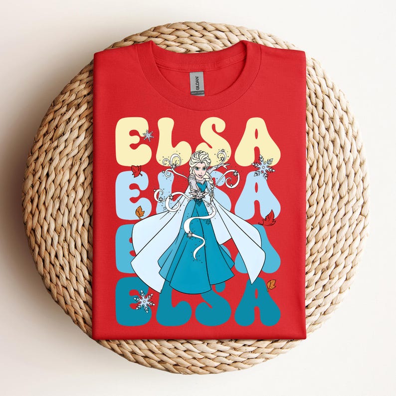 Op de afbeelding: Rood T-shirt met het woord "ELSA" in een retro lettertype, en een afbeelding van Elsa uit Frozen. Het ontwerp bevat sneeuwvlokken en herfstbladeren. Het T-shirt wordt getoond op een geweven, cirkelvormig oppervlak.
