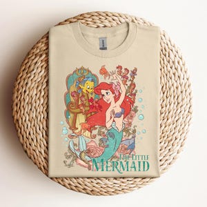 May include: Tan t-shirt featuring a colorful graphic of Ariel from The Little Mermaid. The design includes Ariel with red hair, a blue seashell bra, and a teal tail, surrounded by sea creatures and roses. The words "The Little Mermaid" are printed below.