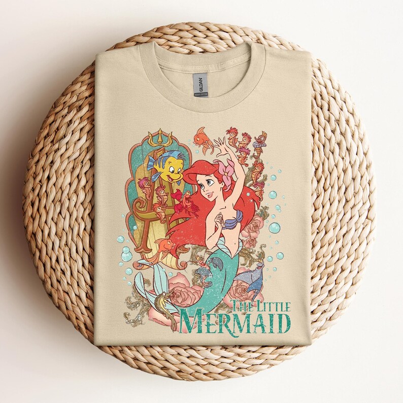 May include: Tan t-shirt featuring a colorful graphic of Ariel from The Little Mermaid. The design includes Ariel with red hair, a blue seashell bra, and a teal tail, surrounded by sea creatures and roses. The words "The Little Mermaid" are printed below.