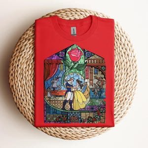 May include: A red t-shirt featuring a stained-glass design with a rose and characters dancing. The graphic includes a couple in yellow and blue attire, with other figures and architectural details. The shirt is displayed on a woven surface.