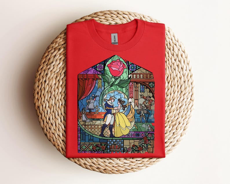 May include: A red t-shirt featuring a stained-glass design with a rose and characters dancing. The graphic includes a couple in yellow and blue attire, with other figures and architectural details. The shirt is displayed on a woven surface.
