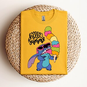 May include: Mustard yellow t-shirt featuring a cartoon character wearing sunglasses and holding a tall ice cream cone. The text "hello summer" is written in a playful font. The shirt is displayed on a woven, circular surface.