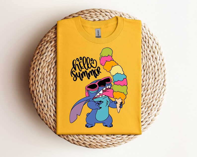 May include: Mustard yellow t-shirt featuring a cartoon character wearing sunglasses and holding a tall ice cream cone. The text "hello summer" is written in a playful font. The shirt is displayed on a woven, circular surface.