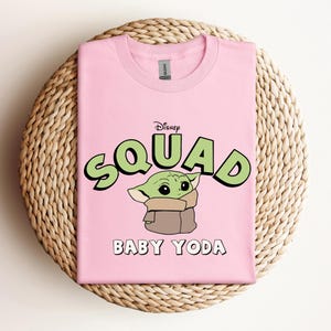 May include: A light pink t-shirt featuring a graphic of Baby Yoda from Disney's The Mandalorian. The word "SQUAD" is in green bubble letters with a black outline, and "BABY YODA" is written below the character in white.