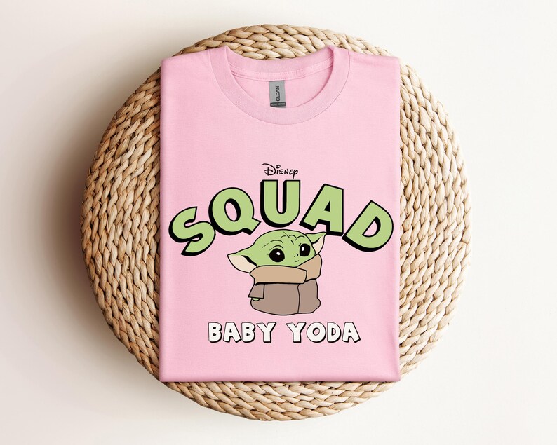 May include: A light pink t-shirt featuring a graphic of Baby Yoda from Disney's The Mandalorian. The word "SQUAD" is in green bubble letters with a black outline, and "BABY YODA" is written below the character in white.