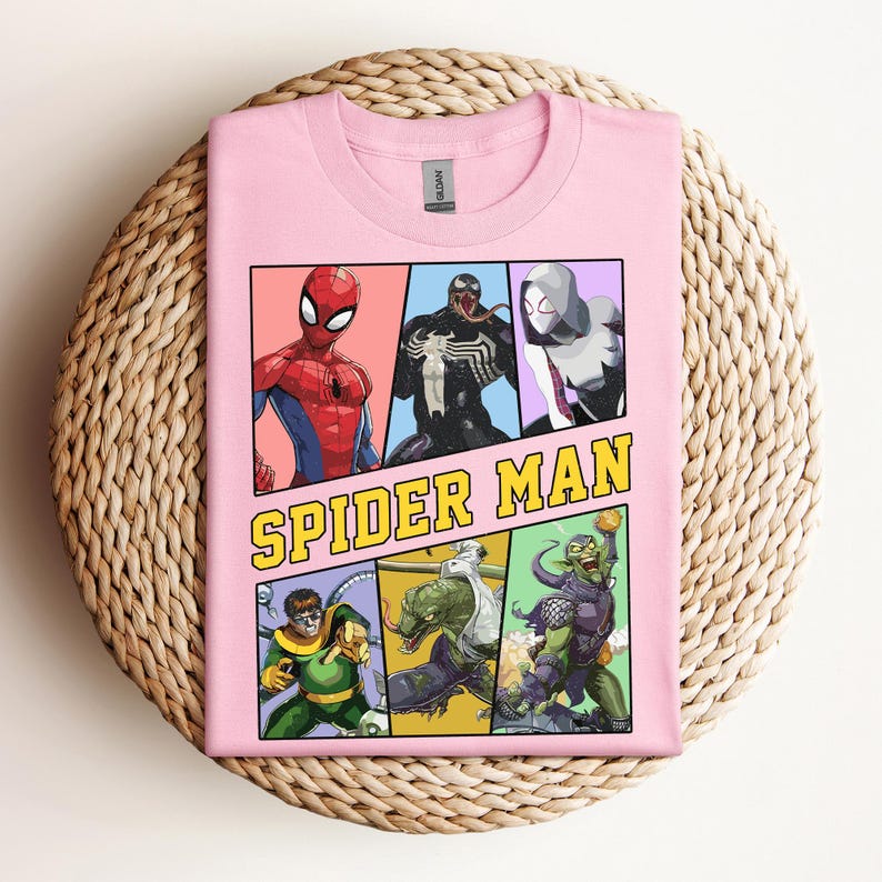 Pode incluir: Uma camiseta rosa claro com um gr&aacute;fico em estilo de hist&oacute;ria em quadrinhos do Homem-Aranha e outros personagens. O gr&aacute;fico inclui o texto "SPIDER MAN" em amarelo e preto. A camisa &eacute; dobrada em uma superf&iacute;cie circular tecida.