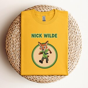 May include: Mustard yellow t-shirt featuring the text "NICK WILDE" above a cartoon image of a fox character in a green circle. The fox is wearing a green shirt and tie. The shirt is folded on a woven, circular surface.