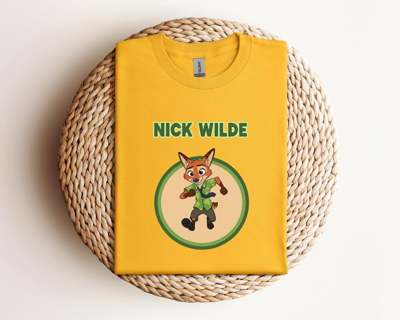 May include: Mustard yellow t-shirt featuring the text "NICK WILDE" above a cartoon image of a fox character in a green circle. The fox is wearing a green shirt and tie. The shirt is folded on a woven, circular surface.