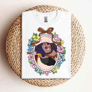 Puede incluir: Camiseta blanca con un dise&ntilde;o de personaje de dibujos animados en un marco ovalado, adornada con flores y una cinta marr&oacute;n. El personaje es morado y negro, sosteniendo un objeto amarillo. La camiseta se muestra sobre una superficie tejida.