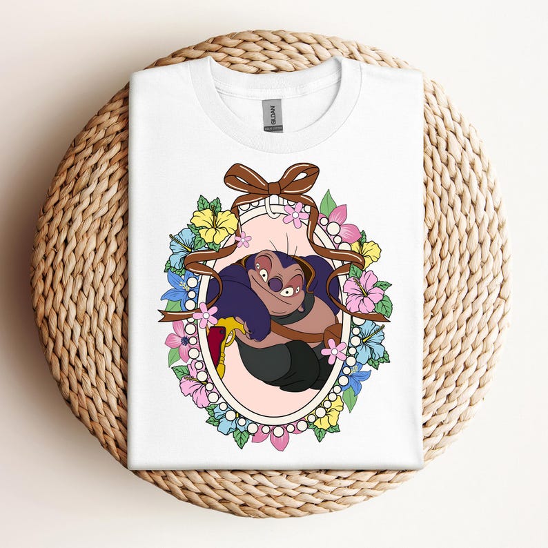 Puede incluir: Camiseta blanca con un dise&ntilde;o de personaje de dibujos animados en un marco ovalado, adornada con flores y una cinta marr&oacute;n. El personaje es morado y negro, sosteniendo un objeto amarillo. La camiseta se muestra sobre una superficie tejida.