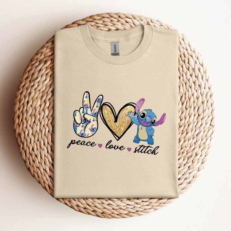 以下が含まれることがあります： ベージュのTシャツに、平和の象徴、ハート、リロ＆スティッチのスティッチが描かれています。平和の象徴は青と紫の花で飾られています。ハートは金色のグリッターです。テキストは「peace love stitch」です。
