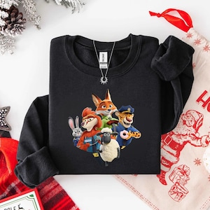 Op de afbeelding: Zwarte crewneck sweatshirt met een kleurrijke afbeelding van personages uit de animatiefilm Zootopia. Het ontwerp bevat een vos, konijn, schaap en andere personages. Een zilveren ketting is zichtbaar.