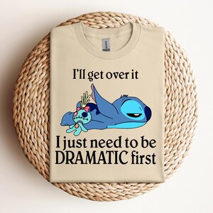 Puede incluir: Camiseta beige con un personaje azul Stitch de Lilo & Stitch. Stitch est&aacute; acostado con una peque&ntilde;a mu&ntilde;eca. El texto en la camiseta dice "I'll get over it I just need to be DRAMATIC first."