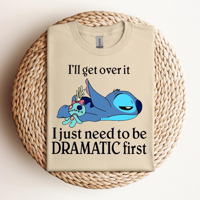 Puede incluir: Camiseta beige con un personaje azul Stitch de Lilo & Stitch. Stitch est&aacute; acostado con una peque&ntilde;a mu&ntilde;eca. El texto en la camiseta dice "I'll get over it I just need to be DRAMATIC first."