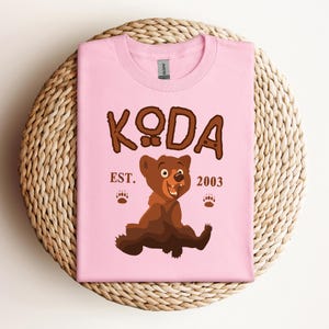 May include: Pink t-shirt with a brown bear cartoon and the text "KODA EST. 2003".