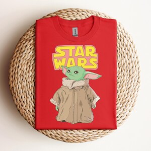 Peut inclure: T-shirt rouge avec un motif d'un personnage extraterrestre vert de Star Wars, avec le texte "STAR WARS" en jaune et rose. L'extraterrestre porte un manteau marron. Le t-shirt est pr&eacute;sent&eacute; sur une surface tiss&eacute;e.